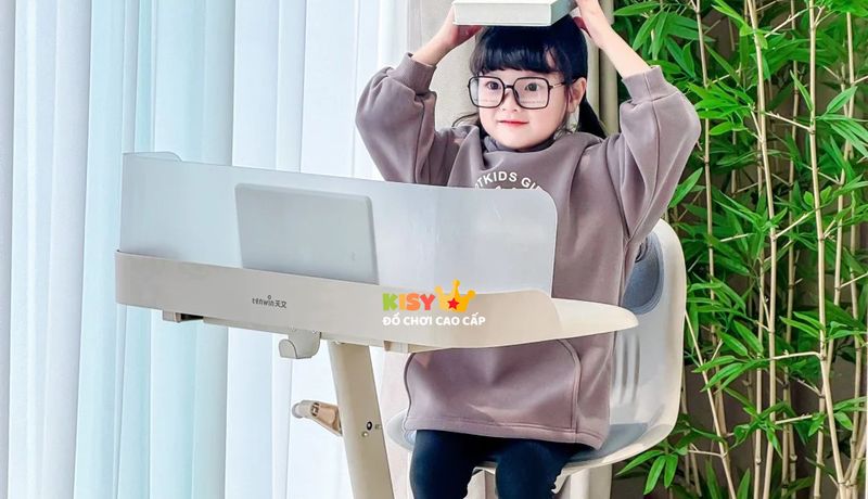Bé ngồi học tại bàn, đội quyển sách trên đầu